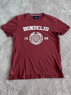 Vinröd Bondelid t-shirt med tryck - Snygg vinröd t-shirt från Bondelid med vitt tryck framtill som visar logga och texten 'BONDELID 1956'. Klassisk rund halsringning och korta ärmar. Tillverkad i mjuk bomull som känns skön mot huden. Perfekt för en avslappnad och stilren look.