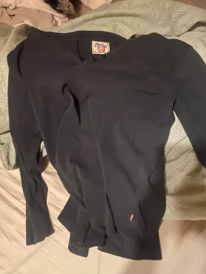 Svart långärmad topp med v-ringning - En svart långärmad topp med v-ringning och enkel design. Tillverkad i mjukt bomullsmaterial, perfekt för en avslappnad och stilren look. Liten röd detalj vid nederkanten ger en extra touch.