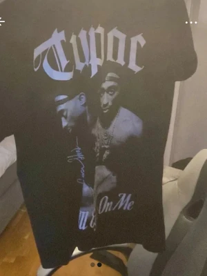Svart Tupac t-shirt med tryck - Säljer en svart t-shirt med stort Tupac-tryck på framsidan. T-shirten har korta ärmar och klassisk passform. Trycket visar Tupac i olika poser och har texten 'Tupac' i stor stil samt autograf-liknande detaljer. Perfekt för dig som gillar hiphop och streetwear.