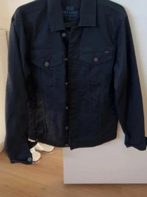 Mörkblå jeansjacka med knappar - Stilren mörkblå jeansjacka med klassisk krage och knappstängning framtill. Två bröstfickor med lock och knapp samt raka ärmar med knapp vid ärmslut. Perfekt att slänga över en hoodie eller t-shirt för en cool look.