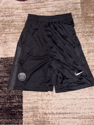 Svarta PSG shorts från Nike - Säljer ett par svarta PSG shorts från Nike med elastisk midja och dragsko. Shortsen har PSG-logga på ena benet och Nike swoosh på det andra. Tillverkade i ett lätt och svalt material som passar perfekt till träning eller sport.