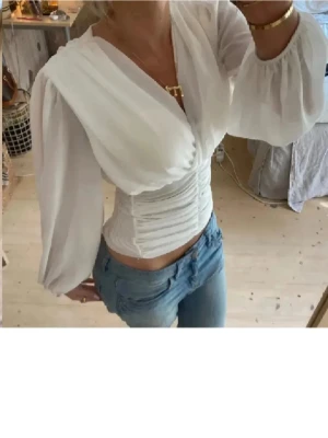 Vit omlottopp med puffärm - Snygg vit omlottopp med markerad midja och långa puffärmar. Toppen har djup v-ringning och draperad effekt över bysten, vilket ger en elegant och trendig look. Perfekt att styla med jeans eller kjol för en chic outfit.