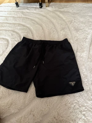 Svarta shorts - Svarta shorts  med elastisk midja och snörning framtill. Klassisk design med diskret Prada-logga på vänster ben. Perfekta för sommaren och tillverkade i ett lätt syntetmaterial som känns svalt och bekvämt.