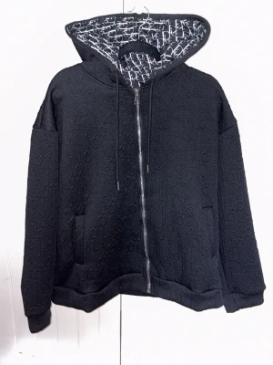 Svart hoodie med dragkedja  - Säljer en svart hoodie med dragkedja och huva. Utsidan har diskret mönster i samma färg och insidan av huvan är fodrad med svartvitt logomönster. Hoodien har fickor framtill och justerbar dragsko i huvan. Perfekt för en trendig och avslappnad look. ((Inte en dior tröja))