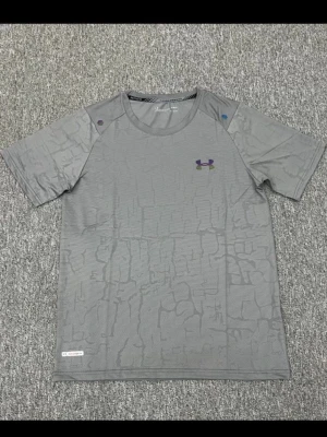 Grå mönstrad t-shirt Under Armour - Snygg grå t-shirt från Under Armour med diskret strukturmönster och korta ärmar. Loggan i lila och gult på bröstet ger en sportig vibe. Perfekt för dig som gillar stilrena träningsplagg.