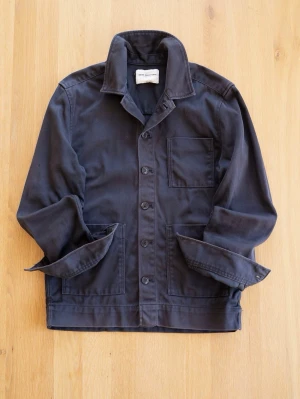 Navy Chore Jacket från These Glory Days - Stilren svart overshirt från These Glory Days med klassisk krage, tre framfickor och knäppning framtill. Tillverkad i ett mjukt bomullstyg som ger en avslappnad men ändå snygg look. Perfekt att bära över en t-shirt eller hoodie.