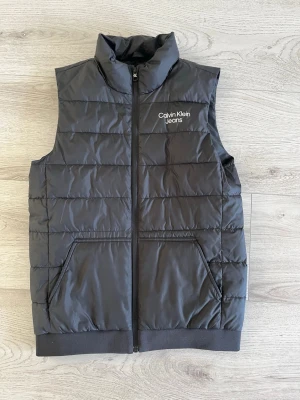 Calvin Klein Väst - Stilren svart dunväst från Calvin Klein Jeans med diskret logga på bröstet. Västen har dragkedja framtill, två fickor och quiltad design. Perfekt för lager-på-lager och ger en clean look till din outfit. Storlek S (170)