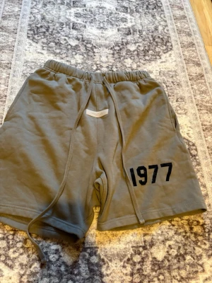 Grå shorts Essentials Fear of God - Säljer ett par grå shorts från Essentials Fear of God med svart '1977'-tryck på ena benet. De har elastisk midja med snörning och är tillverkade i mjuk bomull, perfekta för en avslappnad stil.