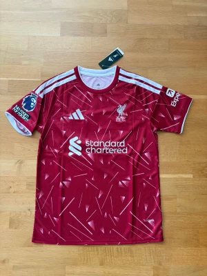 Liverpool matchtröja 26/27 - Snygg röd Liverpool matchtröja från Adidas med vita detaljer och tryck. Tröjan har klubbmärke, sponsorlogga och Premier League-märke på ärmen. Nummer 9 och namnet ISAK på ryggen. Tillverkad i lätt och ventilerande material, perfekt för fotboll eller träning.