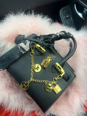 Svart handväska med gulddetaljer - Stilren svart handväska i strukturerat läder med guldfärgade metalldetaljer och dekorativ kedja med berlocker. Väskan har dubbla handtag och en scarf knuten runt handtaget för extra attityd. Perfekt accessoar för dig som gillar lyxig och trendig stil. Storlek 25 