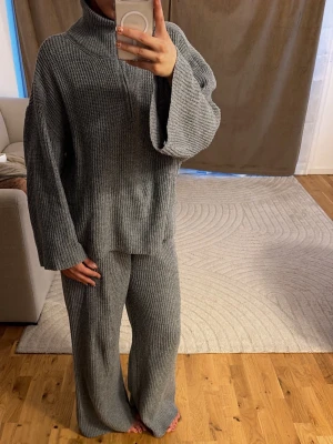 Grått ribbat loungewear-set - Mysigt tvådelat loungewear-set i grå ribbstickad trikå. Setet består av en oversized tröja med polokrage och vida ärmar samt matchande byxor med breda ben. Perfekt för chill dagar hemma eller när du vill vara bekväm men ändå se snygg ut.
