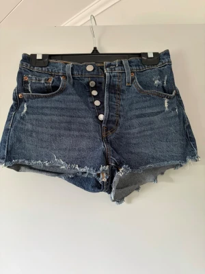 Levi's blå denimshorts hög midja - Säljer ett par blå denimshorts från Levi's med hög midja och råa kanter. Shortsen har knappar framtill, fem fickor och slitna detaljer för en cool vintagekänsla. Perfekta för sommaren och festivaler!