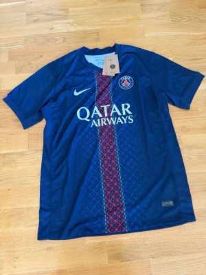 Blå PSG matchtröja från Nike - Snygg blå Paris Saint-Germain matchtröja från Nike med röd och vit grafisk rand i mitten, PSG-logga på bröstet och Qatar Airways-tryck. Tillverkad i lätt och ventilerande material, perfekt för fotboll eller träning. Klassisk rund hals och korta ärmar.