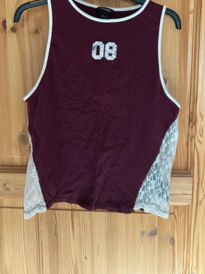 Vinröd linnetopp med spetsdetaljer - Snygg vinröd linnetopp med vita spetsdetaljer på sidorna och vit kant runt hals och ärmhål. Framtill finns en broderad '08'-detalj. Perfekt för dig som vill ha en unik och trendig topp till sommaren.