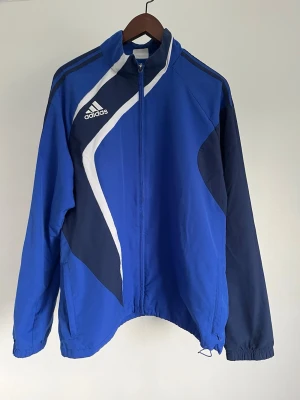 Adidas Teamwear-träningsjacka - Adidas Teamwear-träningsjacka Storlek: XL Passform: Regular Fit  Krage: Hög ståkrage Material: Polyester Mått: Bröstvidd 112 cm, längd 74 cm, axel 50 cm Skick: Träningsjackan är i ett väldigt fint skick utan defekter  Nypris cirka: 800 kr