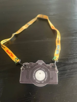 Bad Bunny World Tour kamerahalsband - Unikt halsband med en kameraformad plastbricka och gul textilrem med tryck från Bad Bunny World Tour. Remmen har färgglada texter och detaljer, bland annat 'DeBi TIRAR Más FOTOS' och 'BAD BUNNY'. Perfekt accessoar för fans och samlare.