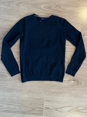 Svart rundhalsad tröja Gant - Klassisk svart tröja från Gant med rund halsringning och långa ärmar. Tröjan har en diskret logga på bröstet och ribbade muddar vid ärmslut och nederkant. Perfekt basplagg för alla tillfällen.