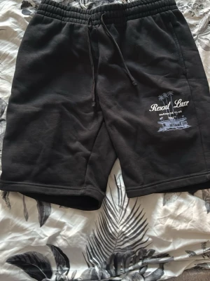 Svarta shorts med tryck och snörning - Svarta shorts med elastisk midja och snörning. Tryck på ena benet med texten 'Resort Luxe Paradise Club' och palmer. Mjukt material, perfekta för sommaren eller chill dagar. Två sidofickor för extra bekvämlighet.