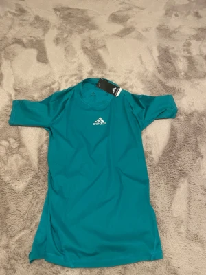Turkos träningströja från Adidas - Snygg turkos träningströja från Adidas med korta ärmar och rund hals. Tillverkad i lätt och ventilerande funktionsmaterial, perfekt för intensiva träningspass. Adidas-logga i vitt på bröstet och diskret meshstruktur för extra komfort.