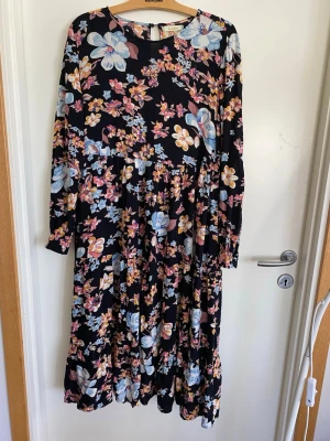 Blommig klänning  med långa ärmar Fire & Glory medium viskos  - Svart klänning  med färgglatt blommönster i blått, rosa och gult. Klänningen har långa ärmar och rund halsringning. Tillverkad i ett mjukt och lätt tyg, perfekt för vår och sommar. Snygg och flowy passform som ger en avslappnad men stilren look.