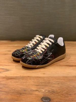 Svarta sneakers med färgstänk - Unika svarta maison margiela sneakers med coola färgstänk i rött, gult, blått och vitt. Skorna har vita snören och en klassisk gummisula i beige. Låg modell med modern streetkänsla och konstnärlig vibe. Perfekta för dig som vill sticka ut med din stil.