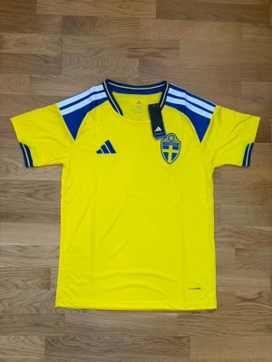 Svensk landslagströja Adidas gul - Gul svensk landslagströja från Adidas med blå detaljer och vita ränder på axlarna. Tröjan har rund hals, korta ärmar och Sverigelogga på bröstet. Tillverkad i lätt och ventilerande material, perfekt för fotboll eller träning.