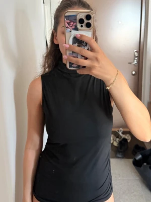 Svart ärmlös polotopp - Säljer en svart ärmlös polotopp med hög hals. Toppen har en slim passform och är tillverkad i ett mjukt, stretchigt material som sitter snyggt på kroppen. Perfekt att styla med jeans eller kjol för en clean look.
