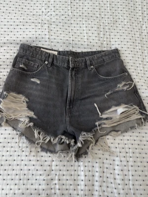 Grå slitna jeansshorts med fransar - Säljer ett par grå denimshorts med hög midja och råa, fransiga kanter. Shortsen har flera slitningar och hål framtill för en edgy look. Klassisk femficksmodell med bälteshällor och dragkedja. Perfekta för sommaren!