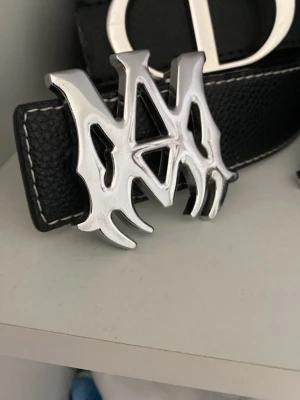 Amiri bälte  - Säljer ett svart bälte i läderimitation med ett stort, silvrigt metallspänne i unik design. Bältet har vita sömmar längs kanterna som ger en snygg kontrast. Perfekt accessoar för att lyfta din outfit.