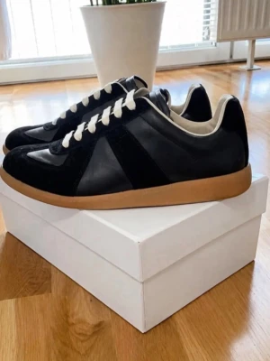 Maison Marigela Gats - Snygga svarta sneakers med beige gummisula och vita snören. Ovandelen är i en mix av mocka och slätt läder, vilket ger en stilren och klassisk look. Insidan är ljus och skon har en låg profil som passar till många olika outfits.