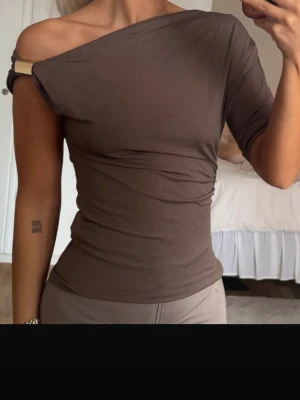 Brun oneshoulder topp från Zara - Säljer en brun oneshoulder topp från Zara med tight passform. Toppen har en unik detalj med metallspänne på ena axeln och är tillverkad i mjukt, stretchigt material. Perfekt för dig som vill ha en trendig och stilren look.