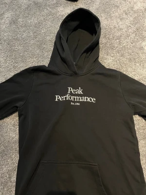 Svart hoodie Peak Performance - Säljer en svart hoodie från Peak Performance med vit broderad logga och text 'Est. 1986' på bröstet. Klassisk modell med huva och känguruficka framtill. Perfekt för en avslappnad och stilren look.