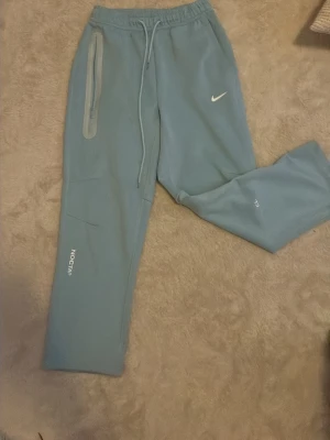 Blå mjukisbyxor från Nike NOCTA - Snygga ljusblå mjukisbyxor från Nike NOCTA med dragsko i midjan och dragkedjeförsedd ficka. Byxorna har tryckta loggor och är gjorda i mjukt material som känns skönt mot huden. Perfekta för chill dagar eller när du vill ha en sportig look.