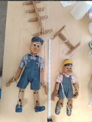 Två trämarionetter i jeanskläder / 2 för 545 kr - Säljer två handgjorda marionettdockor i trä, båda med blå jeansshorts, rutiga skjortor och kepsar. Ena dockan har blå skor och blå keps, den andra har grå skor och gul keps. Dockorna styrs med snören och träpinnar, perfekta för dockteater eller dekoration.