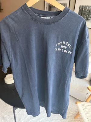 Blå Carhartt t-shirt Class of 89 - Säljer en blå t-shirt från Carhartt med trycket 'Class of 89' på bröstet. T-shirten har rund hals, korta ärmar och är gjord i mjuk bomull. Perfekt för en avslappnad och stilren look.