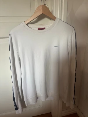 Vit sweatshirt från Hugo - Säljer en stilren vit sweatshirt från Hugo med diskret logga på bröstet och svarta detaljer längs ärmarna. Tröjan har rund halsringning och ribbade muddar. 