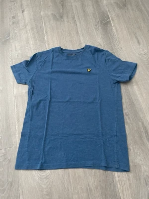Blå t-shirt från Lyle & Scott - En stilren blå t-shirt från Lyle & Scott med klassisk passform och rund hals. T-shirten har korta ärmar och pryds av den ikoniska gula loggan på bröstet. Tillverkad i mjuk bomull som känns skön mot huden.