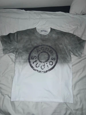 Gråvit t-shirt från Acne Studios - Säljer en snygg t-shirt från Acne Studios med grå och vit färgton och stort tryck på bröstet. T-shirten har korta ärmar och en relaxed fit, perfekt för en trendig streetwear-look. Materialet är mjuk bomull.