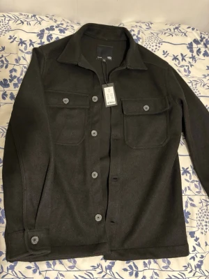 Svart overshirt med fickor NY - Stilren svart overshirt från Lager 157 med klassisk krage och stora bröstfickor med lock. Knäppning framtill med matchande knappar och knappar vid ärmslut. Perfekt lager-på-lager-plagg i ett mjukt och lite tjockare tyg.