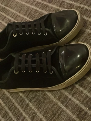 Svarta lanvins - Snygga svarta sneakers med glansig tådel och vita sulor. Skorna har svarta skosnören och metallfärgade öljetter. Klassisk låg modell som passar till många olika stilar. Som ni ser på bilderna är de väll använda! Priset är ej hugget i en sten!
