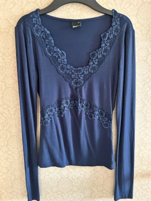 Mörkblå topp med spets från Gina Tricot - Mörkblå långärmad topp från Gina Tricot med dekorativa spetsdetaljer längs ringningen och tvärs över bröstet.  Stretchig men kroppsligpassform. BARA ANVÄNT 1 eller 2 gånger