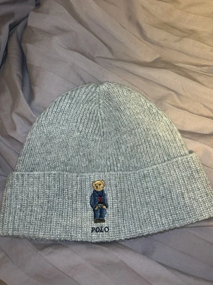 Grå ribbstickad mössa Polo Ralph Lauren - Snygg grå ribbstickad mössa från Polo Ralph Lauren med broderad nalle på uppviket. Tillverkad i en blandning av akryl, nylon och ull. Perfekt accessoar för att hålla värmen och addera lite stil till din outfit. Använt mössan 1 gång. 