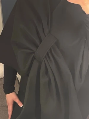 Svart kappa med stor rosett - Säljer en svart kappa med oversized passform och en stor dekorativ rosett framtill. Kappan har vida ärmar och är gjord i ett mjukt tyg som ger ett elegant intryck. Perfekt för dig som vill sticka ut med en unik detalj i din outfit.