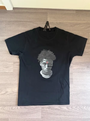 Svart t-shirt med röd V från VLONE - Svart t-shirt från VLONE med stort rött V-tryck på ryggen och ett motiv av ett ansikte med kedja på framsidan. Klassisk rund hals och korta ärmar. Perfekt för dig som gillar streetwear och vill sticka ut med en unik design.