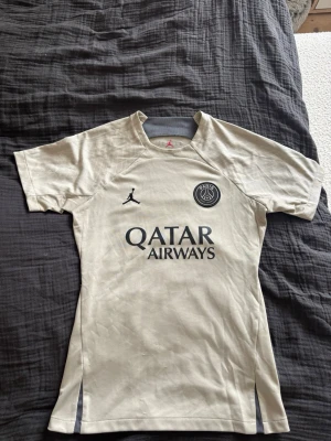 Beige PSG  Jordan träningströja - Snygg beige PSG matchtröja från Jordan med korta ärmar. Tröjan har Paris Saint-Germain-logga och Jordan-logga på bröstet samt Qatar Airways som sponsortryck. Tillverkad i ett lätt och ventilerande material, perfekt för fotboll eller träning. Har lite noppror längst ner se sista bild