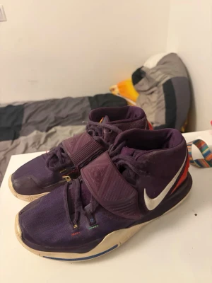 Lila Nike Kyrie 6 sneakers - Säljer ett par lila Nike Kyrie 5 sneakers med vit sula och färgglada detaljer på snörningen. Skorna har en tydlig Nike swoosh i vitt på sidan och kardborreband över vristen. Perfekta för dig som gillar basketstil och vill sticka ut med coola färger.