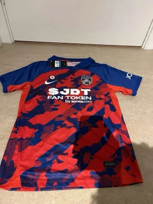 JDT matchtröja Nike röd/blå - Säljer en JDT matchtröja från Nike i rött och blått med kamouflagemönster. Tröjan har klubbmärke på bröstet, sponsortryck och korta ärmar. Tillverkad i lätt och ventilerande material, perfekt för fotboll eller träning.