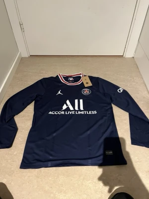 Mörkblå PSG långärmad matchtröja - Snygg mörkblå långärmad matchtröja från PSG i samarbete med Jordan. Tröjan har vita och röda detaljer i kragen, PSG-logga på bröstet och Jordan-logga. Tillverkad i ett lätt och funktionellt material, perfekt för fotboll eller träning.