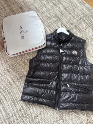 Svart dunväst från Moncler - Stilren svart dunväst från Moncler med hög krage och två dragkedjeförsedda fickor framtill. Västen har Moncler-logga på bröstet och quiltad design. Materialet är glansigt och lätt, perfekt för lager-på-lager och coola outfits. I mycket gott skick size 3