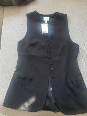 Svart väst med knappar från H&M - Stilren svart väst från H&M med klassisk v-ringning och sex knappar framtill. Västen har två fickor och är tillverkad i ett slätt material som ger en dressad look. Perfekt att styla över en skjorta eller t-shirt🤩